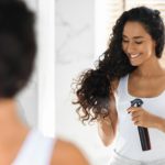 Haircare. Beautiful Young Lady Applying Hair Spray, Standing Near Mirror In Bathroom